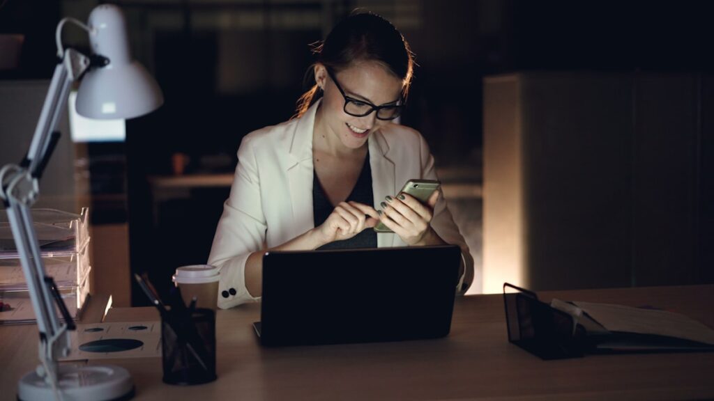 Woman working on phone and laptop sending email at night